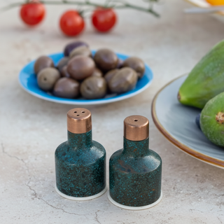 Two blue speckled salt and pepper shakers on a textured surface with olives and avocados in the background.